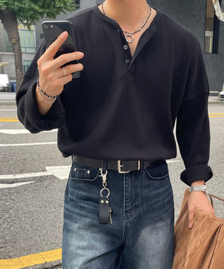 黒のワッフル生地の長袖ヘンリーネックTシャツを着用し、ダークブルーのデニムパンツにタックインしてスマートフォンを操作する男性のアップ。