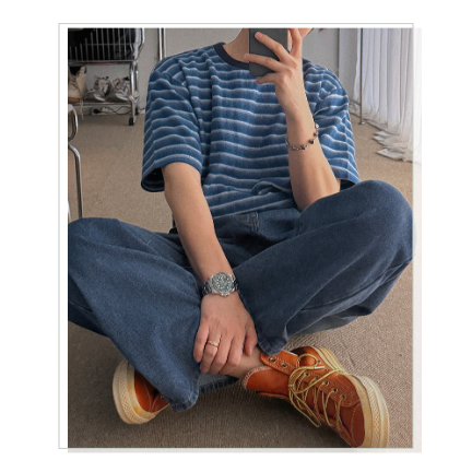 ブルーのストライプTシャツにワイドデニムとオレンジのスニーカーを合わせ、床に座って鏡越しに自撮りする男性の全身カット