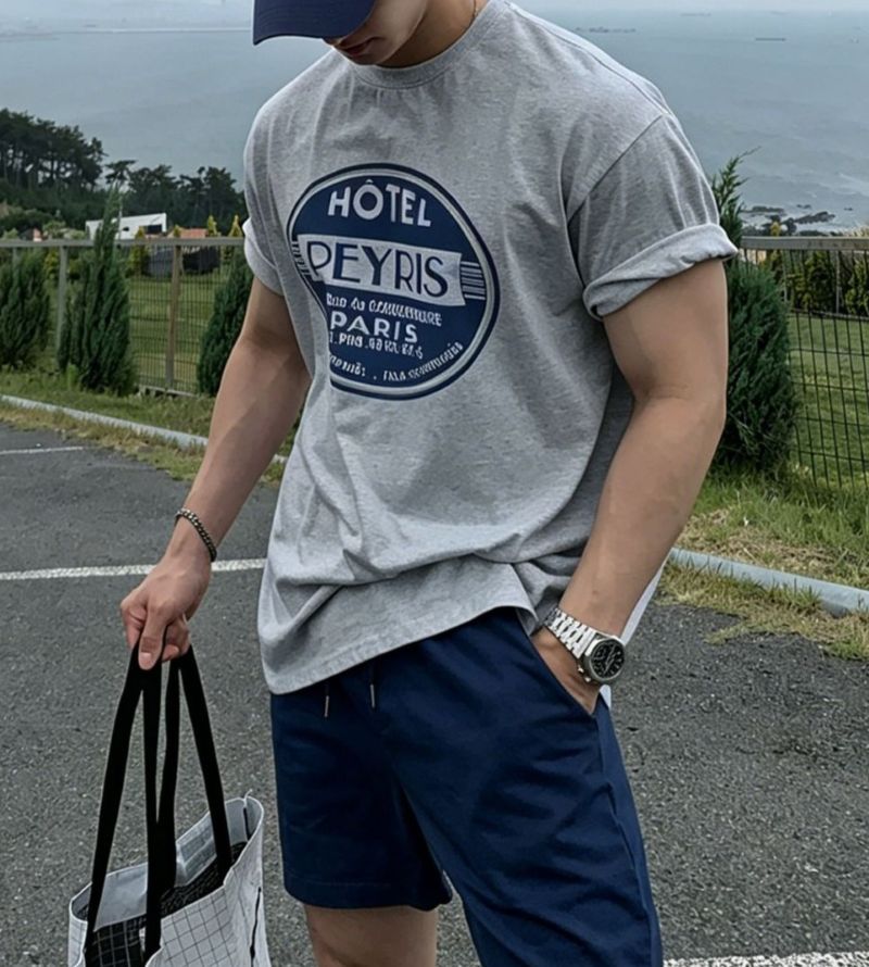 ライトグレーのグラフィックTシャツにネイビーのショートパンツを合わせた男性が、トートバッグを片手に持って歩くスタイリッシュなフロントカット。