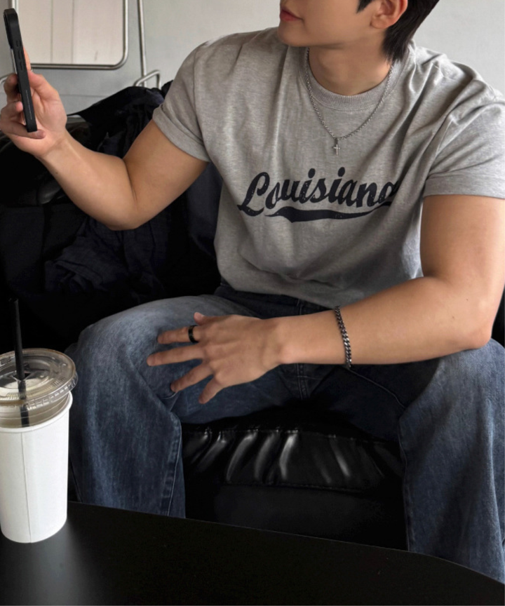グレーのLouisianaロゴ入り半袖Tシャツとウォッシュ加工のデニムパンツを着用し、黒のソファに座ってスマートフォンを操作する男性の斜め前からのクローズアップ。
