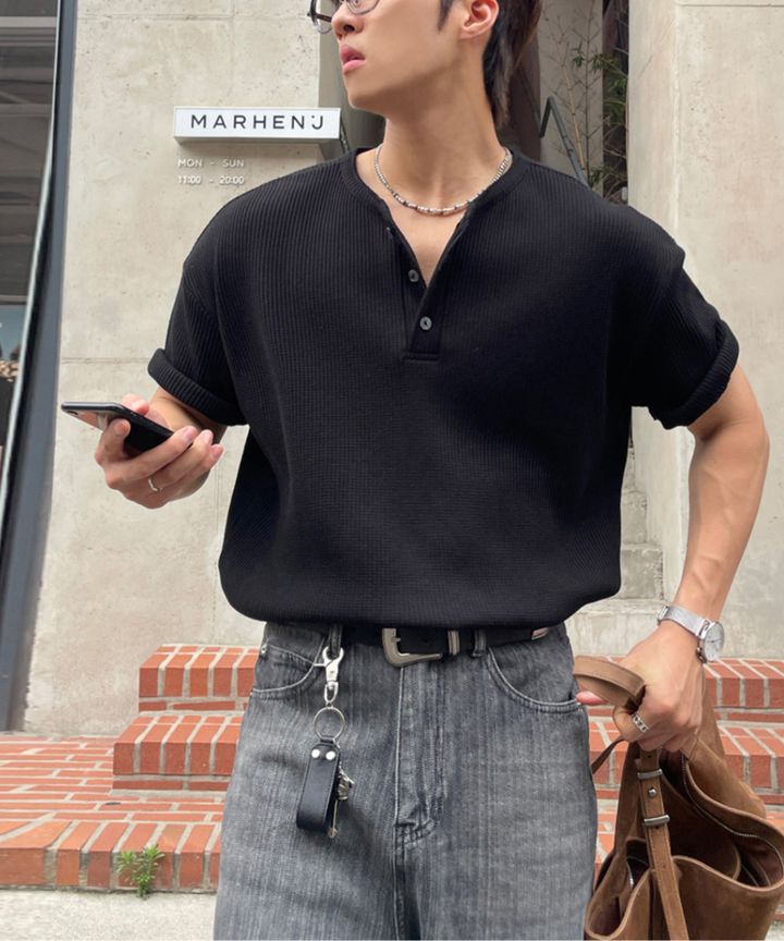 黒のワッフル半袖ヘンリーネックTシャツを着用し、ポケットに手を入れて佇む男性の左側面からのカット