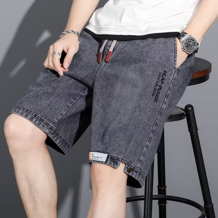 ウォッシュ加工が施されたグレーのメンズデニムハーフパンツを着用し、スツールに座っている男性の斜めからのクローズアップ写真。