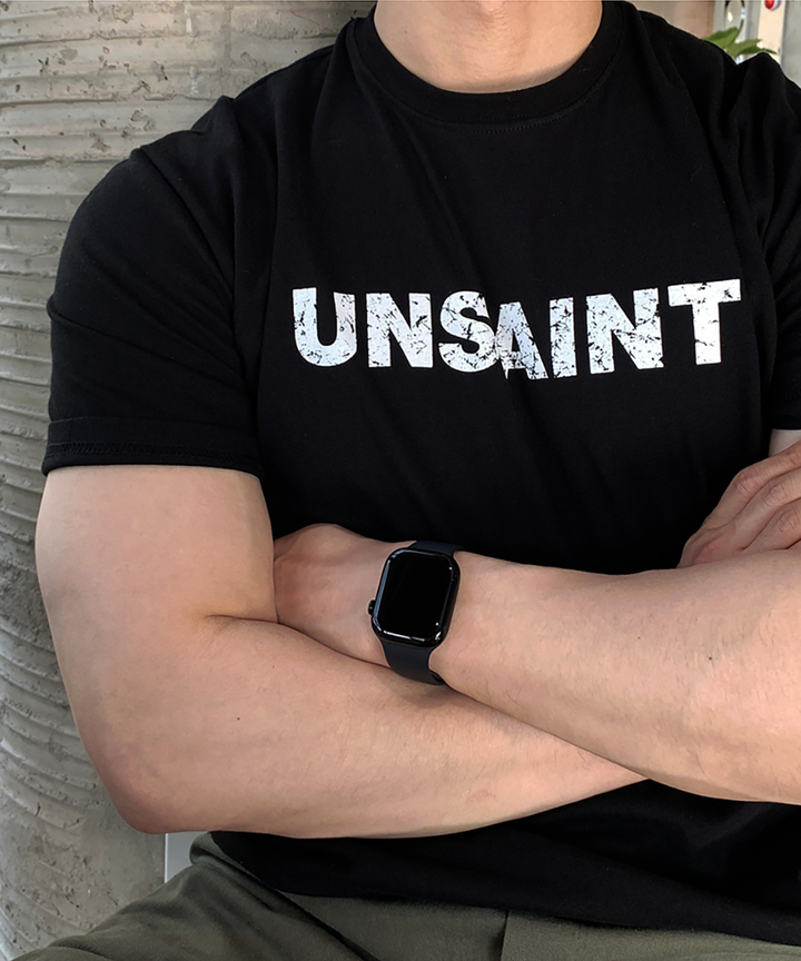 ブラックのボディに白いかすれプリントの「UNSAINT」ロゴが入った半袖Tシャツを着用し、腕を組んだ男性のアップ写真。