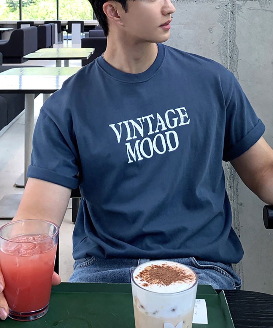 ブルーのVINTAGE MOODロゴTシャツを着用し、カフェの席でドリンクを前にして横を向いている男性の姿