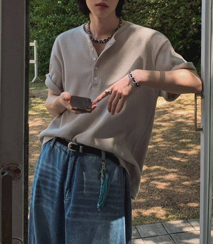 カーキのヘンリーネックワッフルTシャツを着用し、屋外のドア付近でスマートフォンを操作している人物のコーディネート写真。