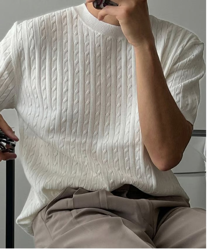 ホワイトの半袖ケーブルニットを着用し、椅子に座る男性の斜め前からのアップ写真
