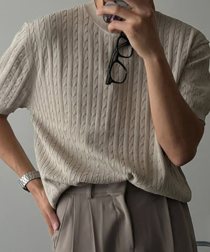 カーキの半袖ケーブルニットを着用し、眼鏡を手元に持ってポーズをとる男性のアップ写真