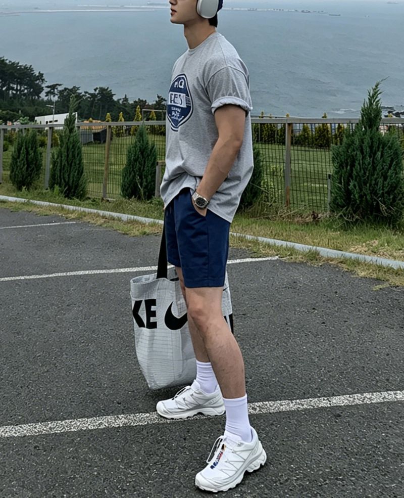 ライトグレーのグラフィックTシャツにネイビーのショートパンツ、白のソックスと白のスニーカーを合わせた男性の全身サイドカット。袖を綺麗にロールアップし、大きなロゴ入りトートバッグを片手に持って海沿いの駐車場に立っています。