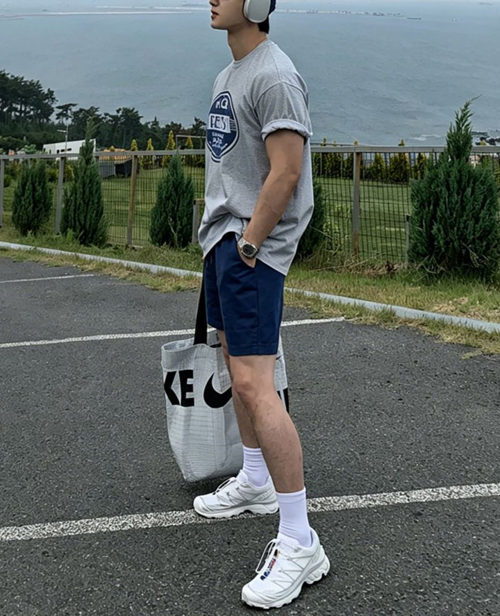 ライトグレーのグラフィックTシャツにネイビーのショートパンツ、白のソックスと白のスニーカーを合わせた男性の全身サイドカット。袖を綺麗にロールアップし、大きなロゴ入りトートバッグを片手に持って海沿いの駐車場に立っています。
