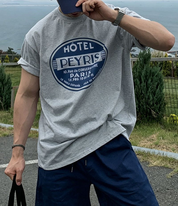 ライトグレーのグラフィックTシャツを着た男性が、右手でネイビーのキャップのツバを軽く掴み、顔を少し傾けているフロントカット。袖はラフにロールアップされており、男らしくスポーティーな雰囲気を演出しています。