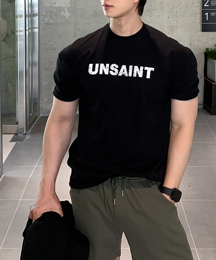 ブラックの「UNSAINT」ロゴTシャツとオリーブのドローコード付きナイロンショーツを着用し、黒いスマートウォッチと黒い折りたたんだシャツを持った男性の全身写真。