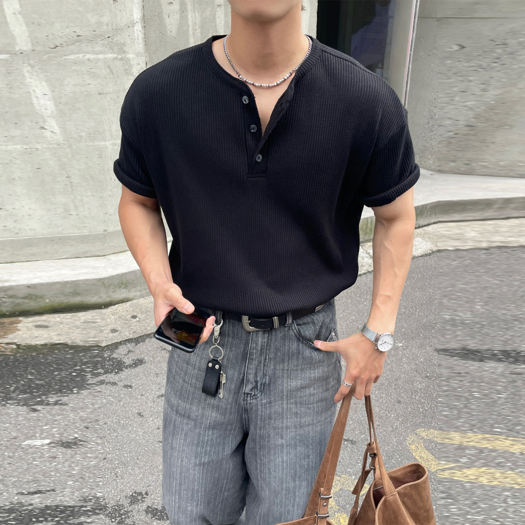 黒の半袖ヘンリーネックTシャツにグレーのワイドデニムパンツと黒のローファーを合わせ、ブラウンのスエード調トートバッグを持った男性の街中での全身コーディネート