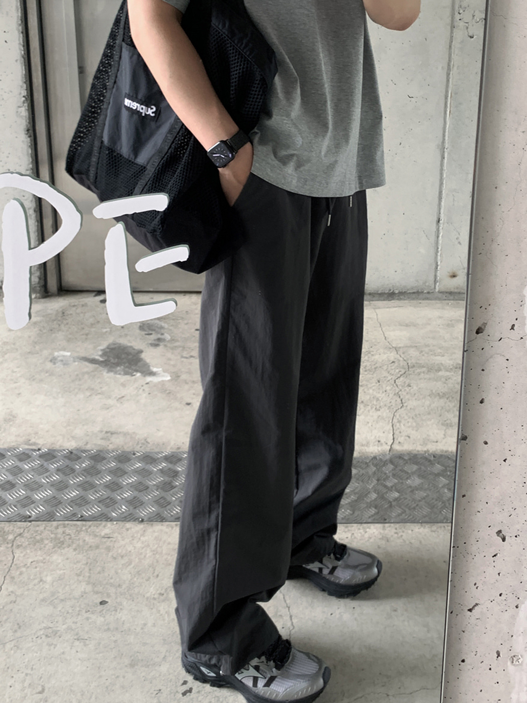 グレーのTシャツにアイアングレーのワイドパンツを着用し、黒いメッシュバッグを肩にかけた男性の鏡越しのサイドシルエット。