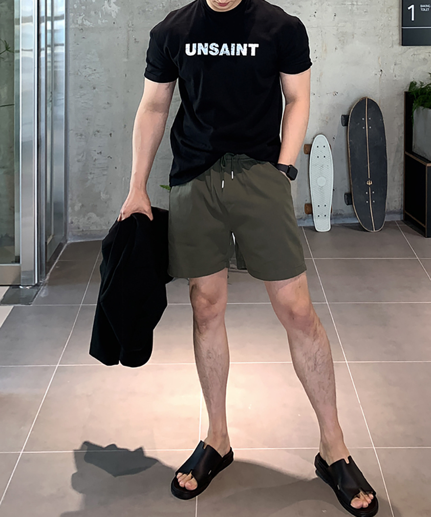 ブラックのロゴTシャツとオリーブのショーツ、黒いレザーサンダルを着用した男性の全身写真。