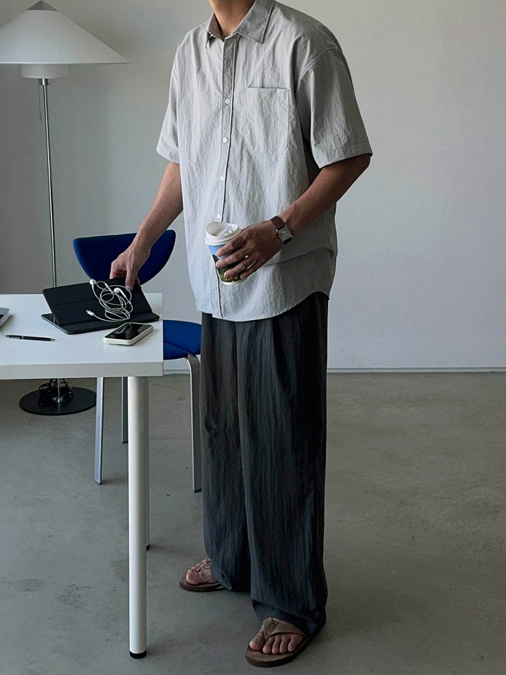 グレーの半袖シャツとダークグレーのワイドパンツを合わせ、コーヒーカップを持って立つ男性の全身カット