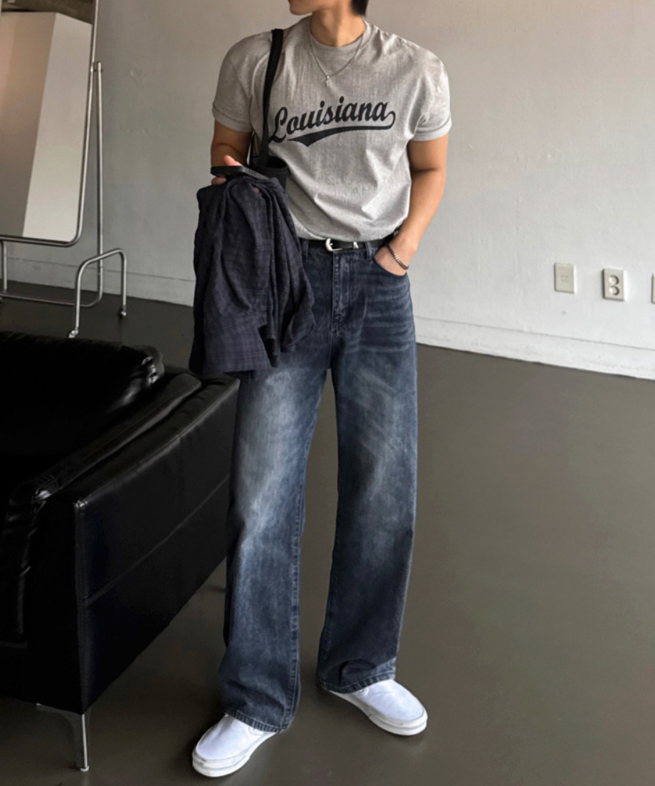 グレーのLouisianaロゴ入りTシャツをデニムパンツにタックインし、黒のベルトを締め、白いスリッポンスニーカーを履いて立つ男性の全身コーディネート。