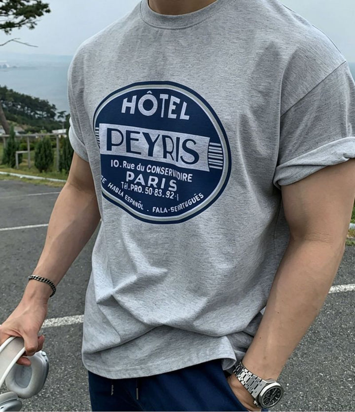 ライトグレーのグラフィックTシャツを着用した男性のフロントアップカット。袖をロールアップし、左腕には存在感のあるメタルバンドの腕時計、右手にはヘッドホンを持っています。