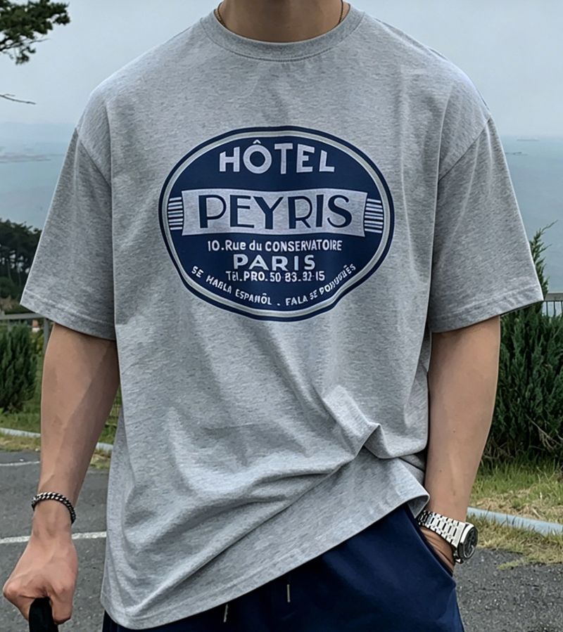 ライトグレーのグラフィックTシャツを着用した男性のフロントアップカット。袖はロールアップせずそのまま下ろしたリラックス感のあるシルエットで、フロントの丸型「HÔTEL PEYRIS」ロゴが中央にはっきりと写っています。