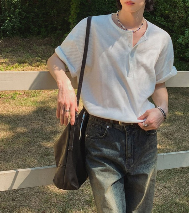 ホワイトのヘンリーネックワッフルTシャツを着用し、木製の柵に寄りかかりながら黒のバッグを肩にかけた人物のウエストアップ写真。