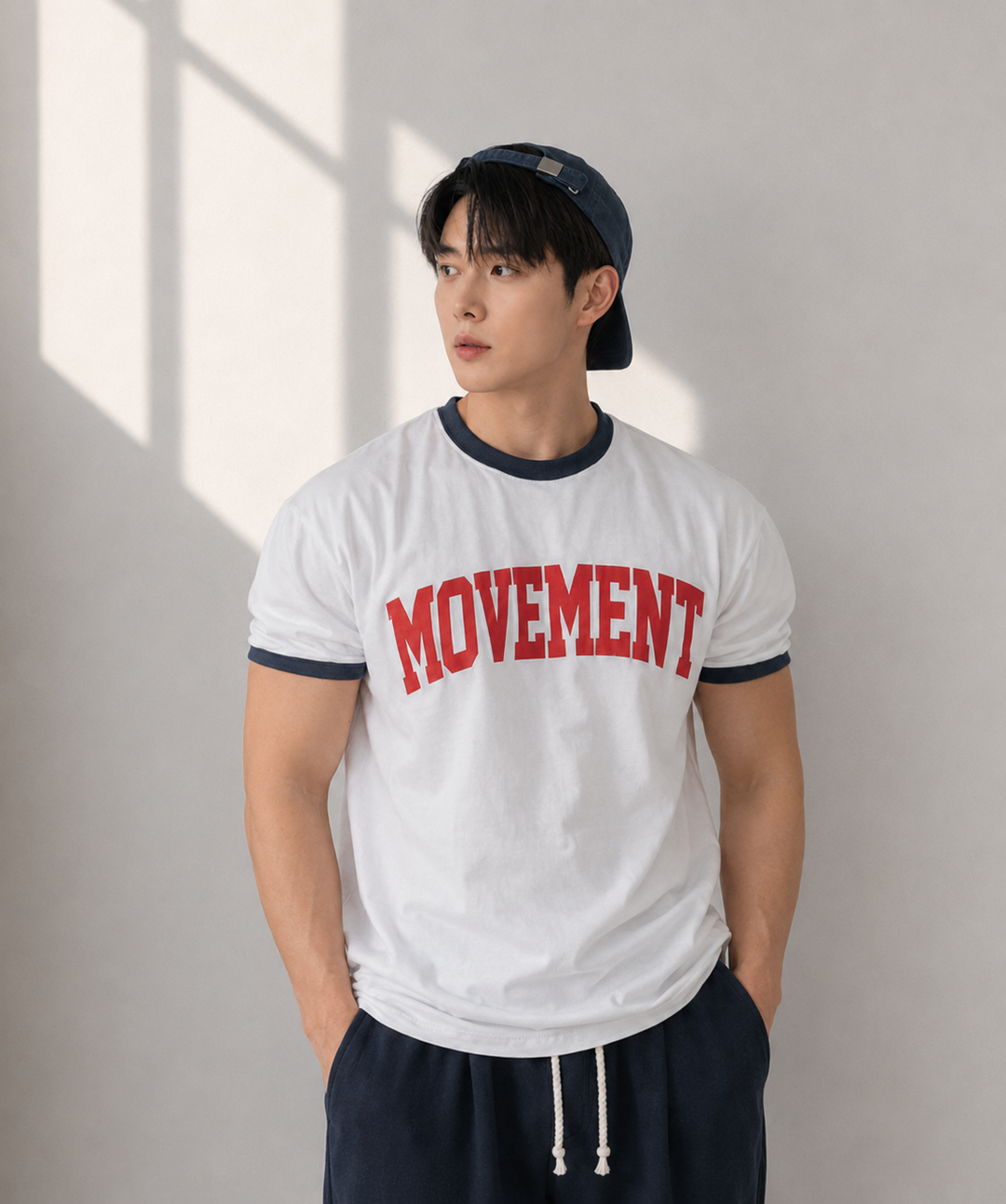 オートミール地に赤のリブ、ネイビーのロゴが入ったリンガーTシャツを着用し、グレーのスウェットパンツのポケットに手を入れた男性の正面アップカット。