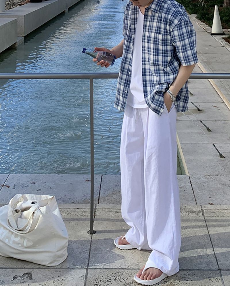 ブルーチェック柄の半袖シャツのフロントを開け、白のワイドパンツに手を入れて立つ男性の全身正面カット。左手には青いボトルのミネラルウォーターを持っています。