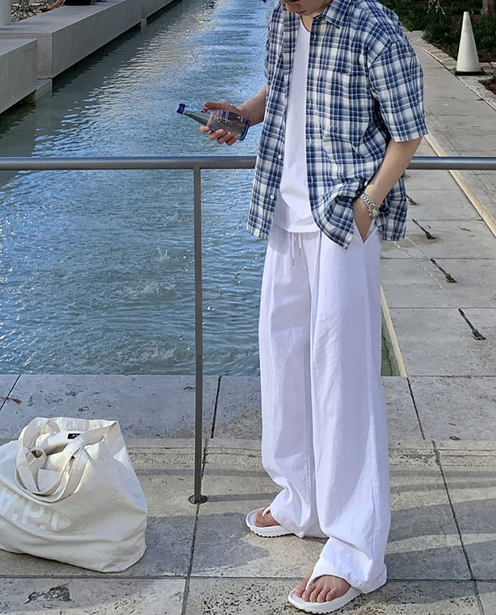 ブルーチェック柄の半袖シャツのフロントを開け、白のワイドパンツに手を入れて立つ男性の全身正面カット。左手には青いボトルのミネラルウォーターを持っています。