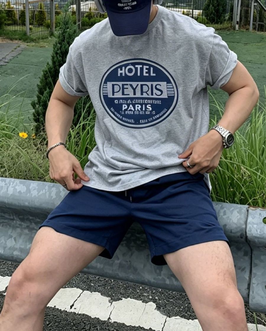 ライトグレーのグラフィックTシャツの裾を両手で軽く整えながら、ガードレールに腰掛ける男性のポーズカット。ネイビーのショートパンツや小物とのバランスが際立つ夏のアウトドアスタイルです。