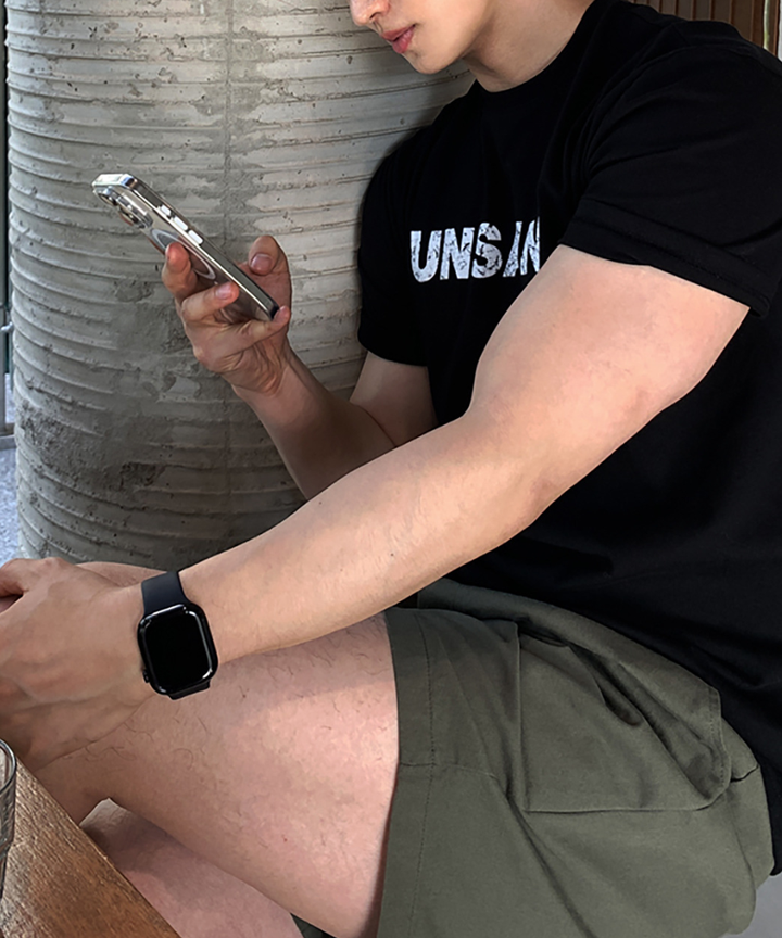 ブラックのロゴTシャツとオリーブのショーツを着用し、座りながらスマートフォンを操作する男性の横顔。