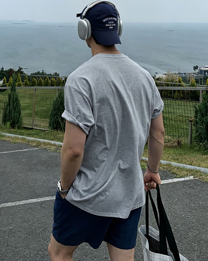 ライトグレーのグラフィックTシャツにネイビーのショートパンツ、ネイビーのキャップを後ろ被りし、ヘッドホンを装着した男性のバックスタイル。右手には大きめのトートバッグを持っています。