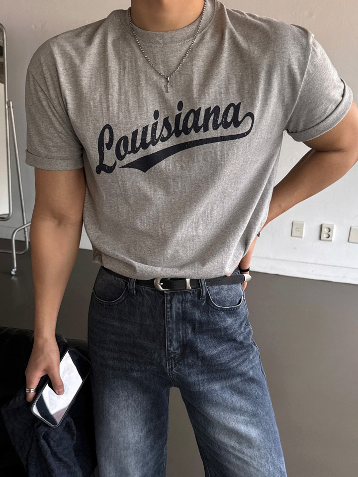 グレーのLouisianaロゴ入り半袖Tシャツを着用し、左手を腰に当てて立つ男性の上半身スタイリングカット。