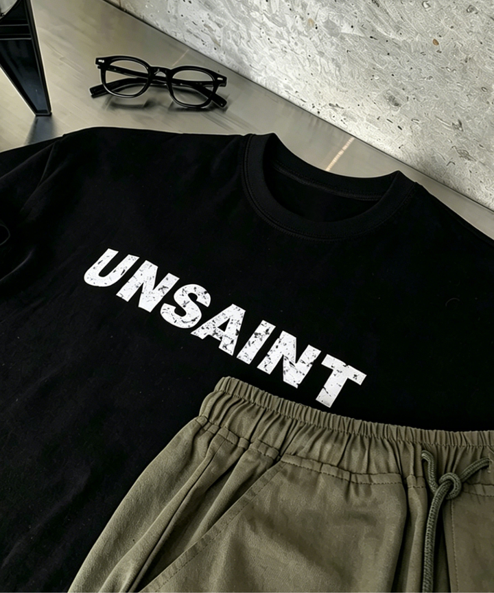 ブラックの「UNSAINT」ロゴTシャツ、オリーブのナイロンショーツ、黒縁メガネ、コンクリート調の背景。