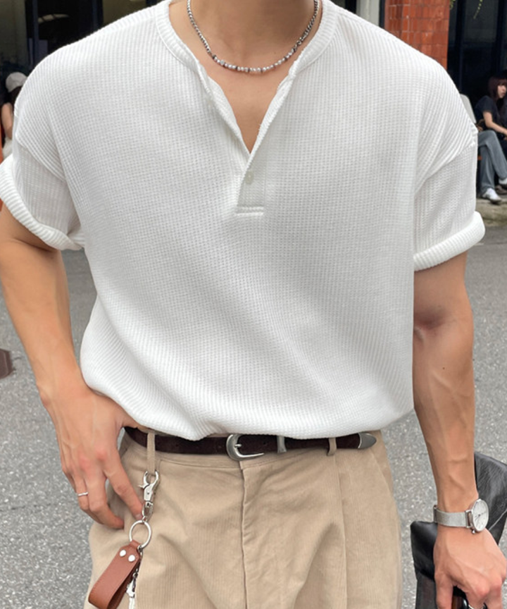白の半袖ヘンリーネックTシャツにベージュのチノパンツを合わせ、黒のレザーバッグを手にした男性の屋外でのコーディネート