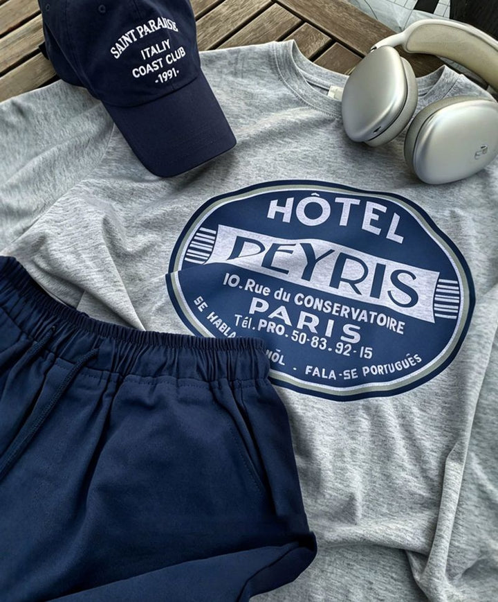 木製テーブルの上に、ライトグレーのグラフィックTシャツ、ネイビーのショートパンツ、ネイビーのロゴキャップ、シルバーのワイヤレスヘッドホンを綺麗に並べた平置きコーディネートカット。
