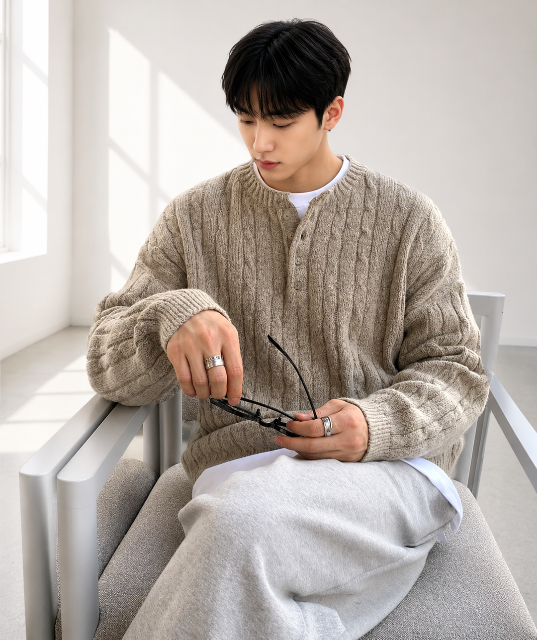 ベージュのケーブルニットを着用した男性が、手元に黒いメガネを持ちながら椅子に腰掛けている落ち着いた雰囲気のクローズアップカット。