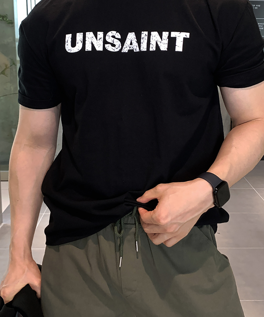 ブラックの「UNSAINT」ロゴTシャツとオリーブのショーツを着用し、スマートウォッチをつけた手でTシャツの裾を整える男性。