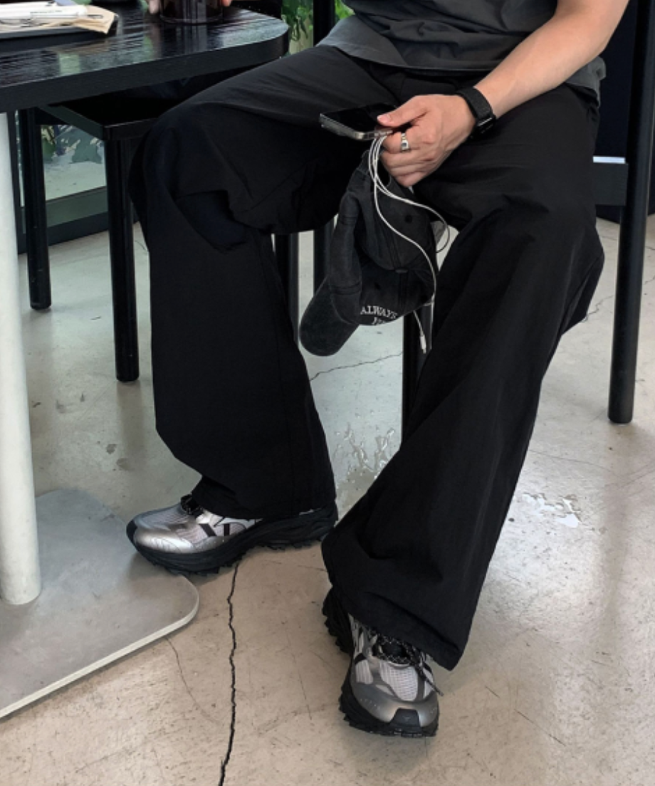 ブラックのナイロンパンツとシルバーのスニーカーを履いて、椅子に座りながらスマートフォンを持つ男性の足元の写真。