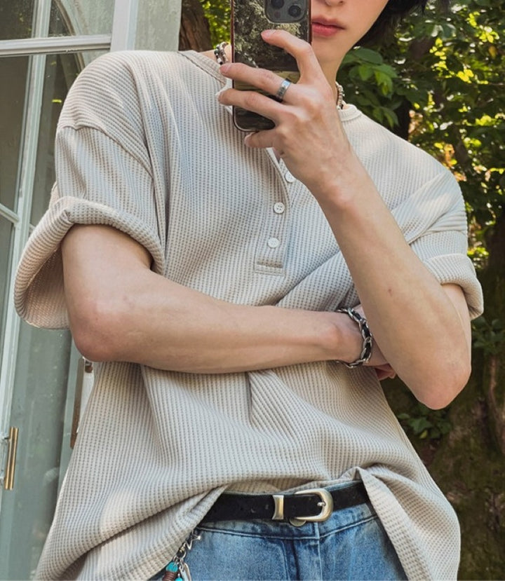 カーキのヘンリーネックワッフルTシャツを着用し、腕を組んで鏡越しにスマートフォンで自撮りをする人物のバストアップ写真。