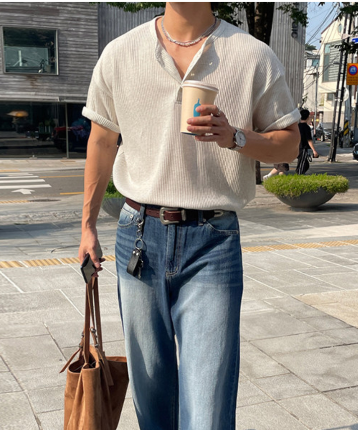 アイボリーのワッフル生地の半袖ヘンリーネックTシャツを着用し、ブルーのウォッシュ加工デニムパンツにタックインした男性の屋外でのアップ。