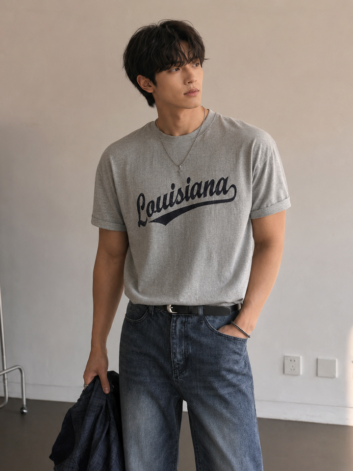 グレーのLouisianaロゴ入り半袖Tシャツをウォッシュ加工のデニムパンツにタックインし、黒のベルトを締めて立つ男性の上半身正面カット。