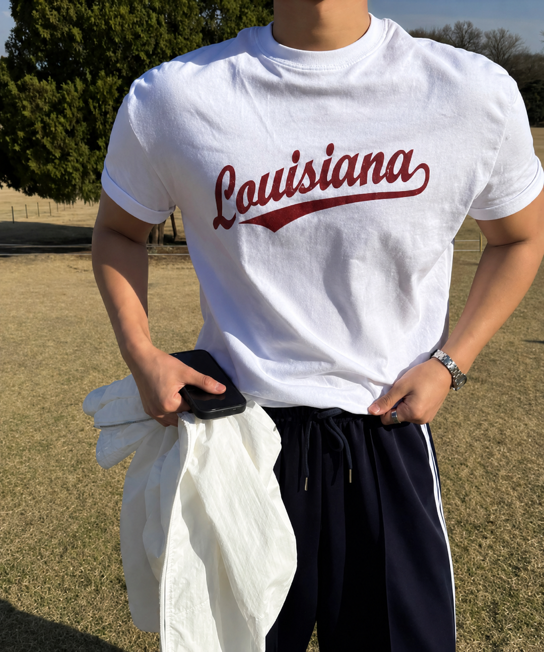 ホワイトのLouisianaロゴ入り半袖Tシャツの裾を整えながら、白いアウターとスマートフォンを手にする男性の屋外での上半身ショット。