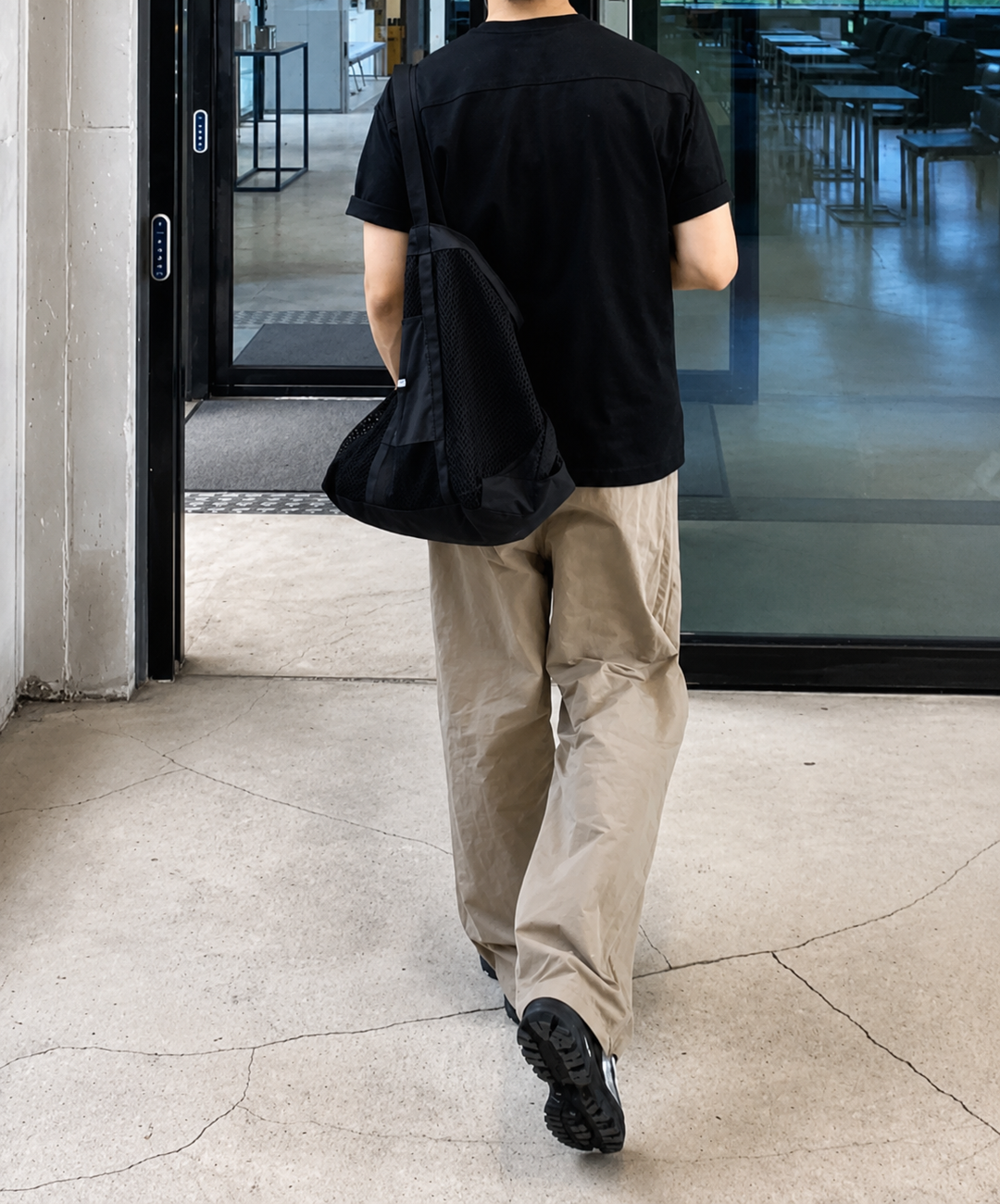 黒のTシャツにベージュのワイドナイロンパンツを履いて、黒のメッシュバッグを肩に掛けた男性の後ろ姿の歩行写真。