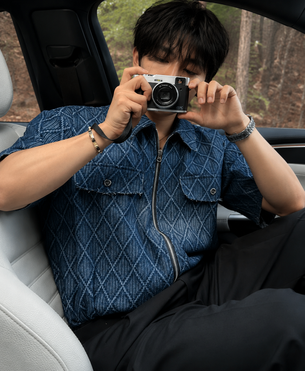 インディゴブルーのハーフジップ半袖シャツに黒いパンツを着用し、車のシートに座る男性のアップカット。