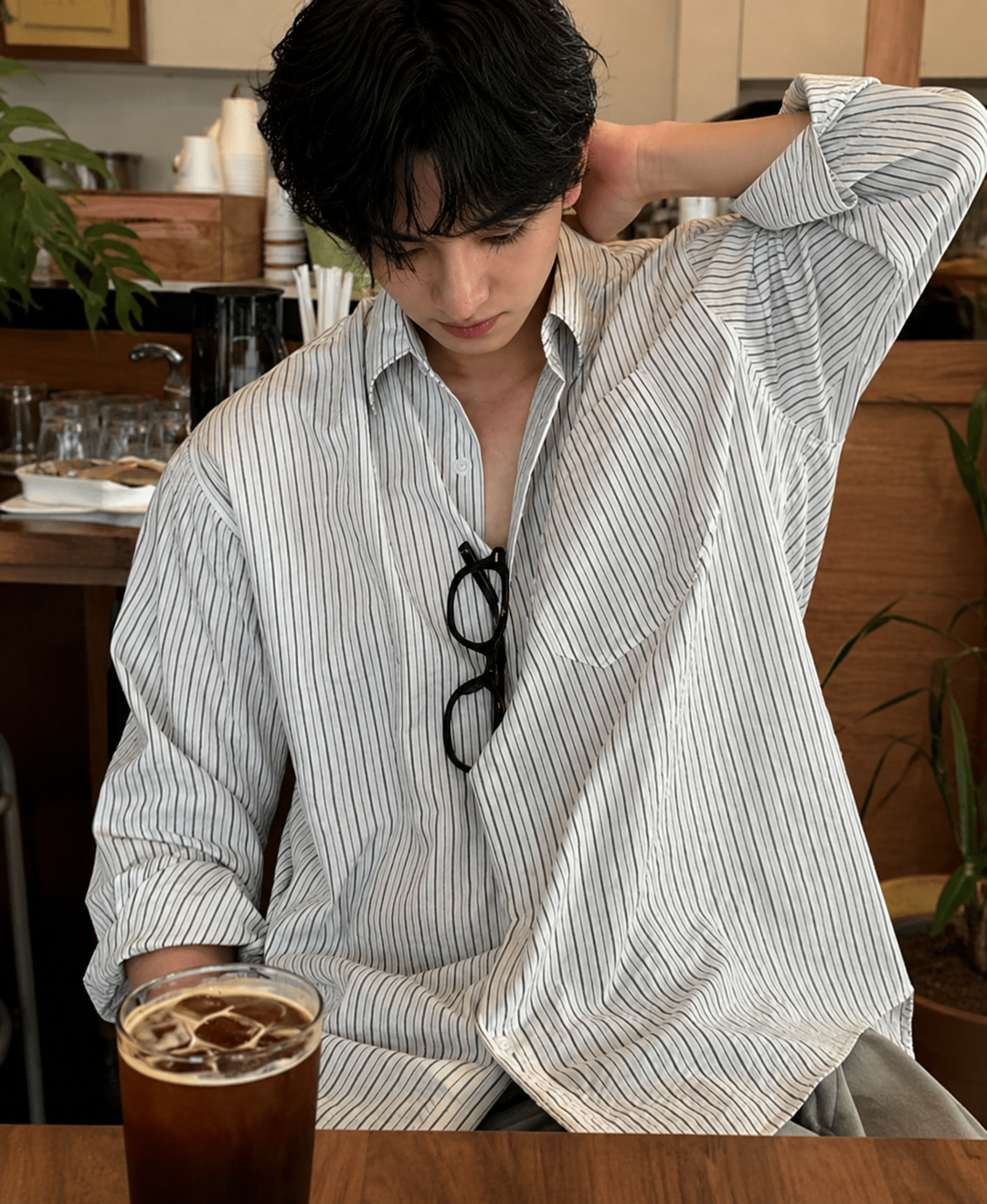 カフェのテーブル席で、ホワイト地に黒の細ストライプが入ったリラックスフィットの長袖シャツを着用し、アイスコーヒーを前に男性の胸元アップ。シャツの胸ポケットや、襟元に掛けられた黒縁のメガネが写っています。