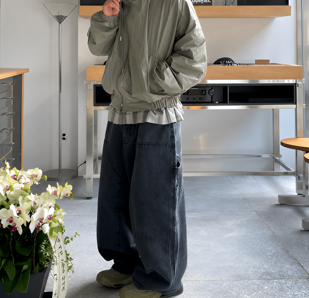 カーキのブルゾンとブラックのワイドパネルデニムパンツを着用し、室内に立っている人物の斜め前からの全身カット。