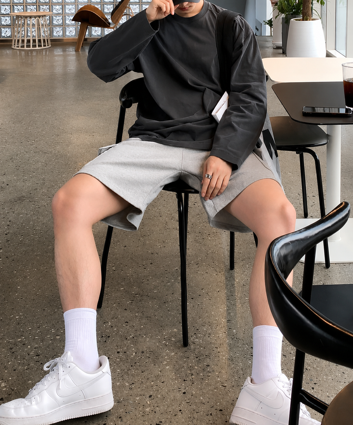 アイアングレーの長袖Tシャツ、ライトグレーのハーフパンツ、白ソックスと白のローカットスニーカー（を合わせた男性が、カフェの椅子に深く腰掛けている全身スタイリングカット。