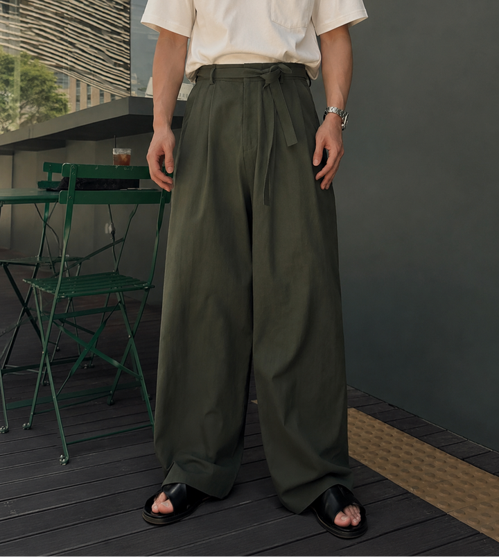カーキのリネンブレンドラップワイドパンツを穿き、足元に黒のレザーサンダルを合わせた男性の立ち姿のアップカット。