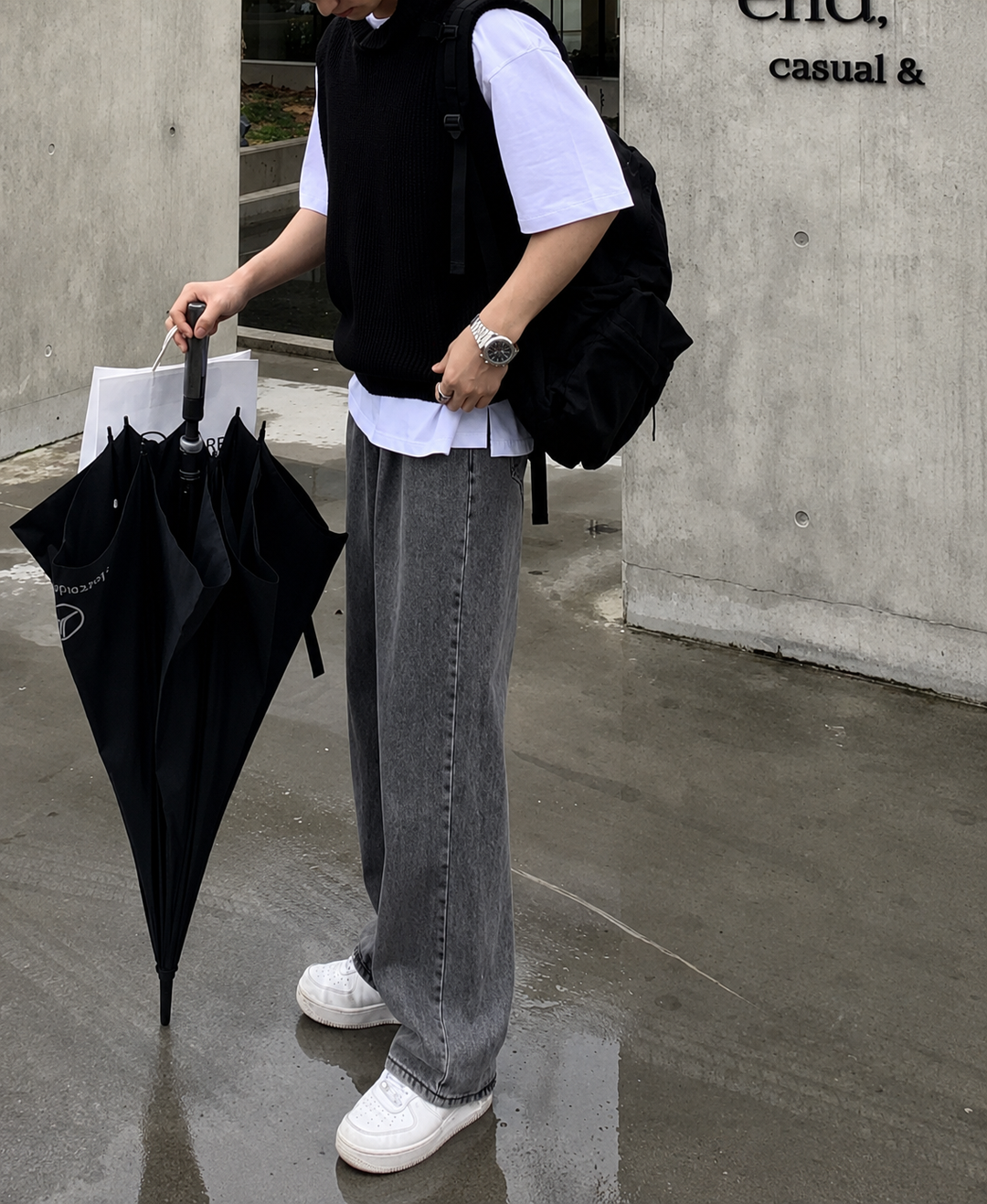 コンクリート壁の建物の前で、白Tシャツにブラックのフード付きニットベスト、グレーのワイドデニムを合わせ、黒いリュックを背負って傘を手に持つ男性のサイド全身ショット