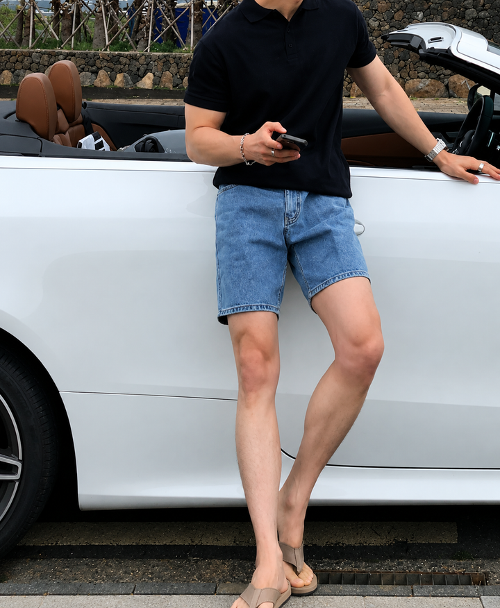 ブラックのポロシャツを着用した男性が、スマートフォンを手に持ちながらオープンカーのドアに寄り添う、リラックスしたリゾートカジュアルのミドルショット。