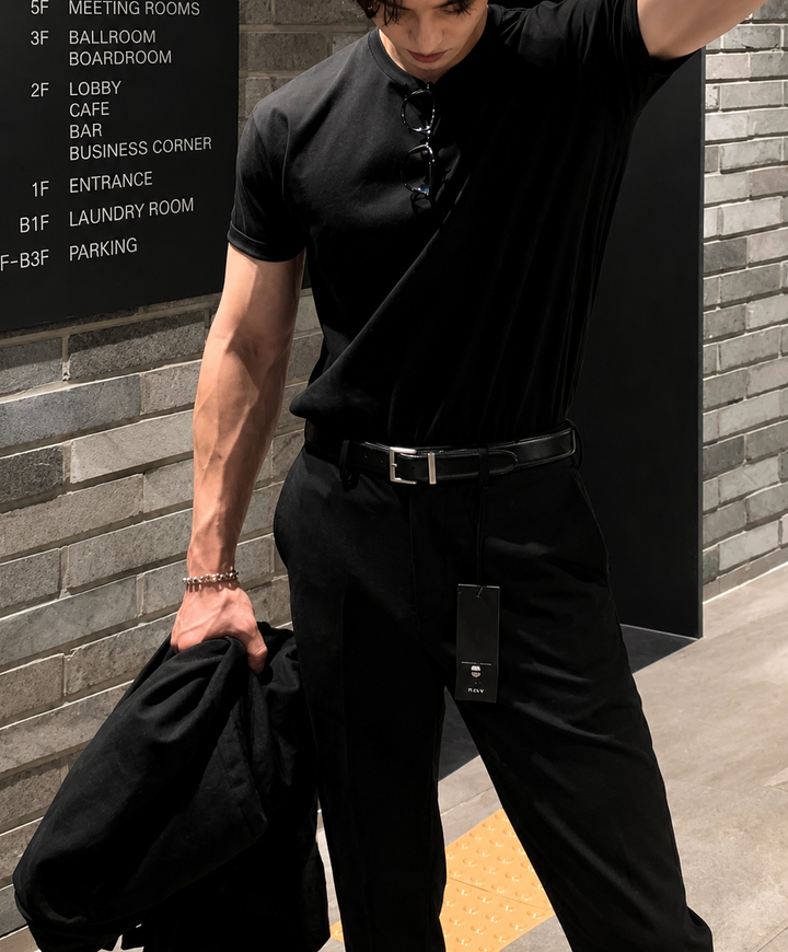 首元にサングラスを掛けたオールブラックコーデ。ブラックのヘンリーネックニットTシャツを黒のベルトとパンツに合わせ、ジャケットを手に持って佇む人物のフロントアップ。