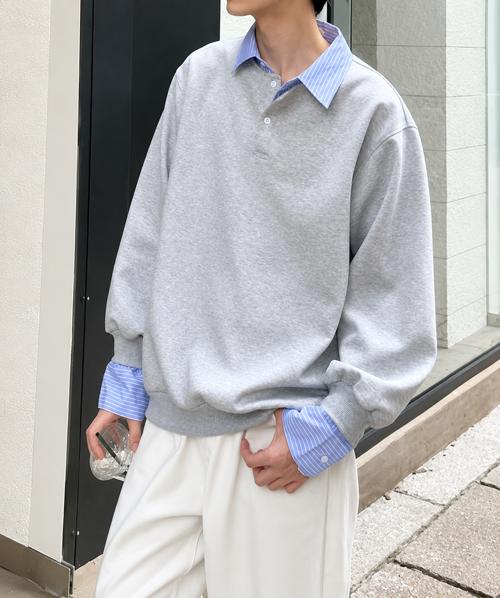 ストライプシャツを重ね着したようなデザインのグレーのポロネックスウェットに、白のワイドパンツを合わせた男性の屋外での立ち姿。